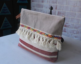 May include: A beige canvas clutch with a zipper closure and a white fringe trim. The fringe is accented with a colorful embroidered band of red, yellow, blue, and green thread.