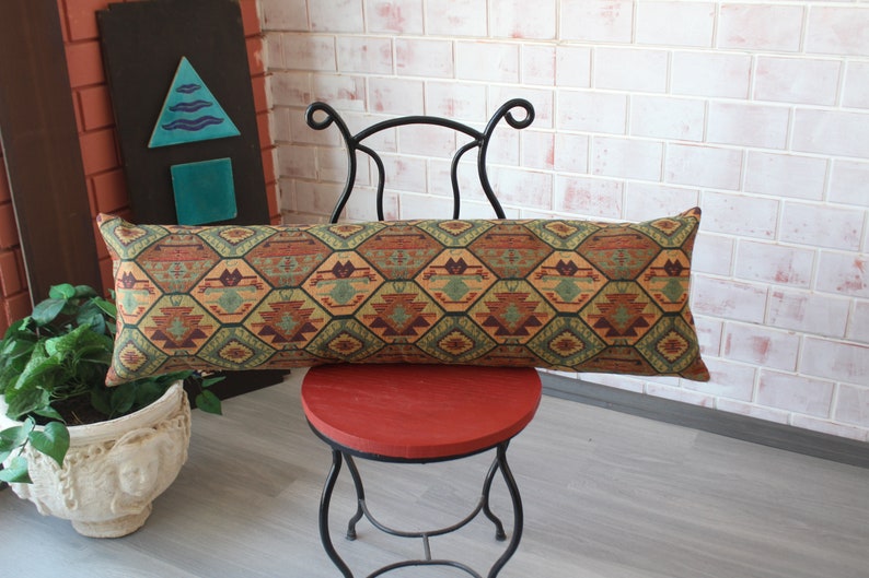 May include: A long, rectangular pillow with a geometric pattern in shades of brown, green, and orange. The pillow is resting on a red chair with a black metal frame.
