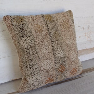 May include: A square decorative pillow with a textured, neutral-toned fabric. The pillow features a repeating pattern of small, off-white and brown shapes. The pillow is leaning against a white wooden surface.