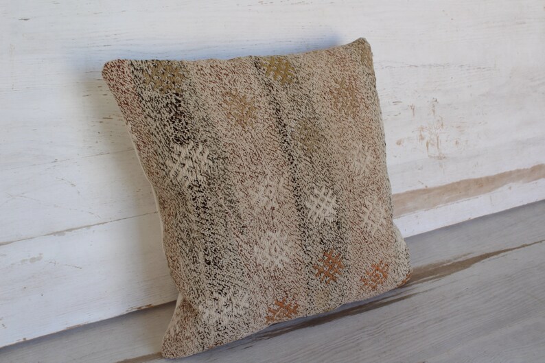 May include: A square decorative pillow with a textured, neutral-toned fabric. The pillow features a repeating pattern of small, off-white and brown shapes. The pillow is leaning against a white wooden surface.