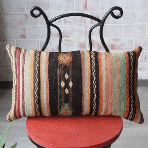 May include: A colorful striped throw pillow with intricate woven patterns in shades of orange, green, brown, and white. The pillow is sitting on a red chair with a black metal frame.