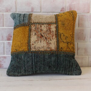 Puede incluir: Una almohada cuadrada con un diseño de patchwork. La almohada está hecha de diferentes telas de colores, incluyendo amarillo, marrón y azul. La almohada está sentada sobre una superficie blanca.