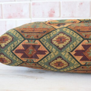 May include: A long, rectangular pillow with a geometric pattern in shades of brown, green, and orange. The pillow is made of a woven fabric with a textured surface.