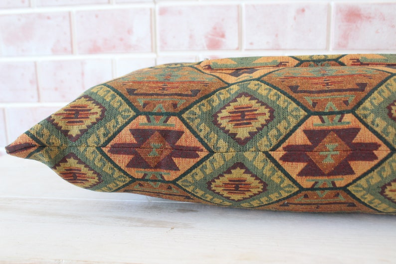 May include: A long, rectangular pillow with a geometric pattern in shades of brown, green, and orange. The pillow is made of a woven fabric with a textured surface.
