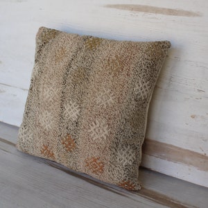 May include: A square decorative pillow with a textured, woven design in shades of beige, brown, and cream. The pillow features a repeating geometric pattern and is leaning against a white wooden surface.