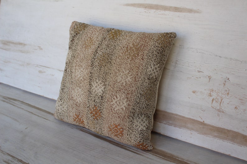 May include: A square decorative pillow with a textured, woven design in shades of beige, brown, and cream. The pillow features a repeating geometric pattern and is leaning against a white wooden surface.
