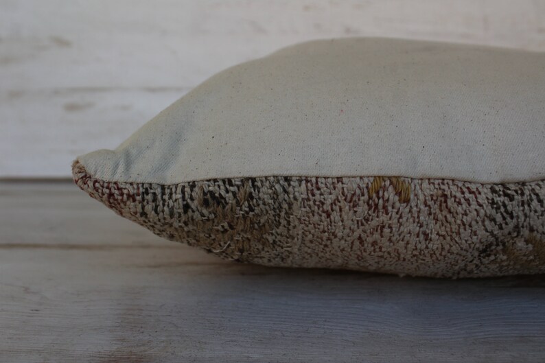 May include: A rectangular pillow with a cream-colored top and a textured, woven bottom. The bottom section features a mix of brown, beige, and red tones, creating a rustic aesthetic. The pillow is designed for home decor.