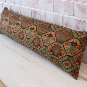 May include: A long, rectangular pillow with a geometric pattern in shades of green, brown, orange, and red. The pillow is made of a woven fabric and has a fringed edge.