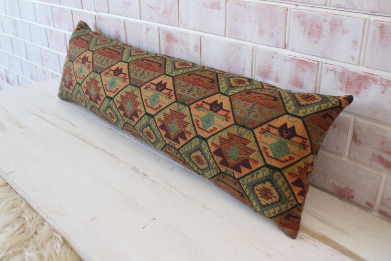May include: A long, rectangular pillow with a geometric pattern in shades of green, brown, orange, and red. The pillow is made of a woven fabric and has a fringed edge.
