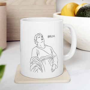 以下が含まれることがあります： 白いセラミックマグカップ。黒い線画で上を見上げる人物と「BRUH」の文字が描かれています。マグカップはベージュのコースターの上に置かれています。