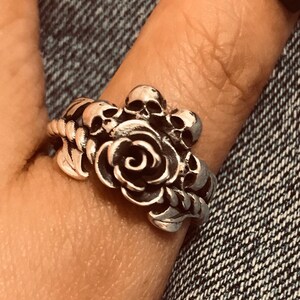 May include: A silver ring featuring a rose design with skull accents. The ring is worn on a finger.