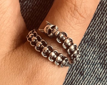 May include: A silver ring with a skull design. The ring is worn on a finger and is made of a chain of small beads.