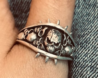 May include: A silver ring with a skull design and spikes around the band. The ring is worn on a person's thumb.