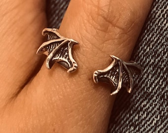 May include: A silver ring with a design of two open wings, worn on a finger with black nail polish.