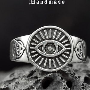 May include: Silver ring with an eye design and a sunburst pattern. The ring is set on a dark gray background. The text "De Me Handmade" is visible above the ring.