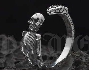 Peut inclure: Une bague en argent représentant un squelette tenant un serpent. La bague est faite à la main et le nom de la marque "De Me" est visible en arrière-plan.