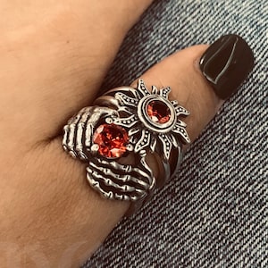 Può includere: Un anello d'argento con un designo dettagliato di due mani che tengono un sole con una pietra preziosa rossa. L'anello è indossato al dito di una persona.
