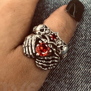 May include: A silver ring with a red gemstone and skull designs. The ring is worn on a person's thumb. The text "De Me Handmade" is visible in the background.
