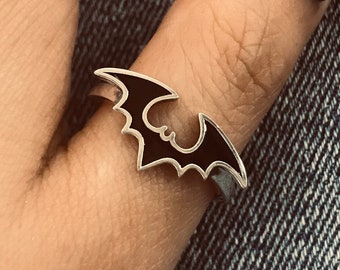 May include: A silver ring with a black bat design. The bat has a black enamel fill and is set against a denim background. The ring is worn on a finger with a dark nail polish.