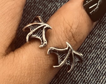 May include: A silver ring with a detailed design of two wings, worn on a finger.