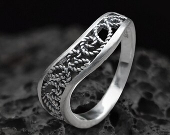 May include: A silver ring featuring a delicate, intricate design. The ring is crafted from a twisted rope pattern and has a slightly curved shape. The ring is displayed against a dark, textured background.