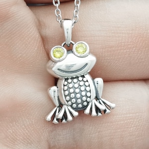May include: A silver chain necklace with a frog pendant. The frog has yellow eyes and a textured body. The necklace is labeled "De Me Handmade".