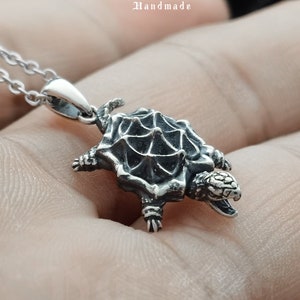 Pode incluir: Um colar com um pingente de prata em forma de tartaruga, com um design de casca texturizado e detalhado. O colar está em uma corrente de prata e está sendo segurado em uma mão. O pingente é rotulado como "De Me Handmade".