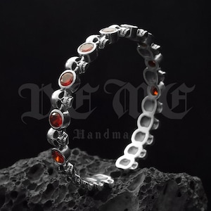 May include: Silver bracelet with red gemstone accents and skull charms. The bracelet is on a dark grey rock. The text "De Me Handmade" is visible in the image.