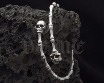 May include: A silver skeleton hand bracelet with two skull charms. The bracelet is handmade and the brand name "De Me" is written in a gothic font above the bracelet.