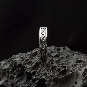 May include: A silver ring with a decorative, swirling design. The ring is sitting on a dark, textured surface.
