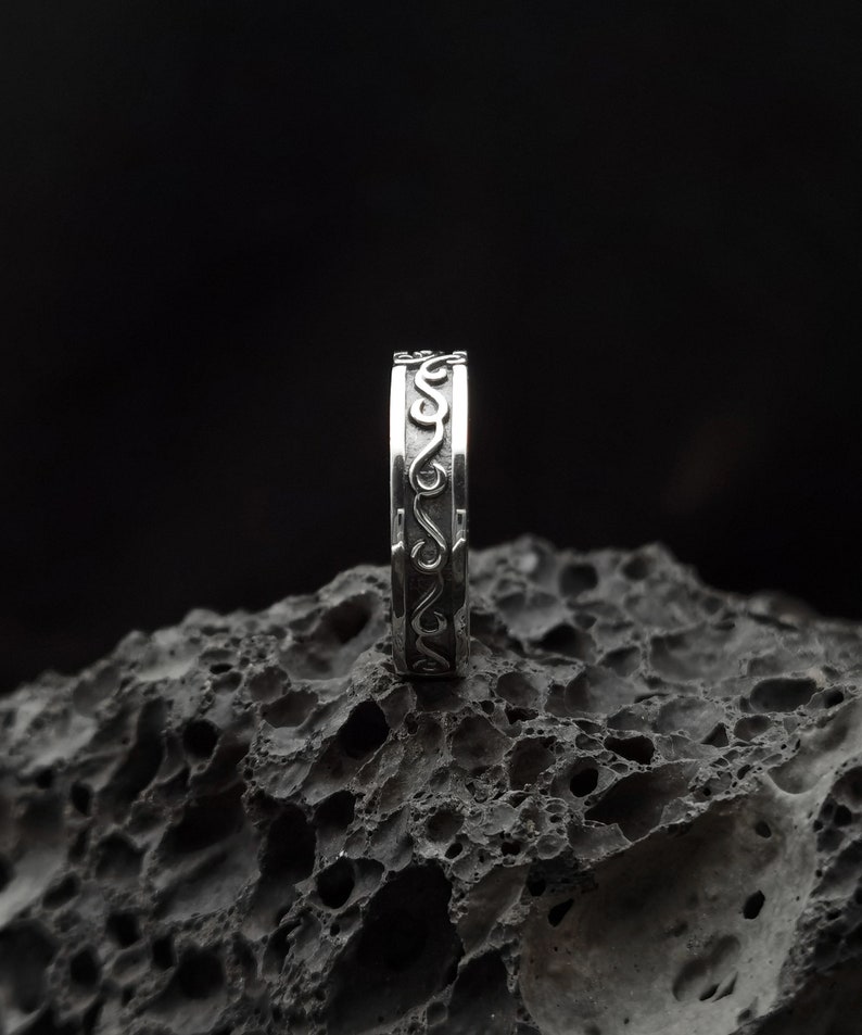 May include: A silver ring with a decorative, swirling design. The ring is sitting on a dark, textured surface.