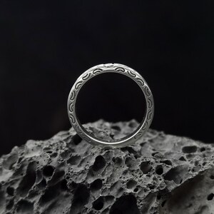 May include: A silver ring with a detailed, intricate design. The ring is sitting on a dark, textured surface.