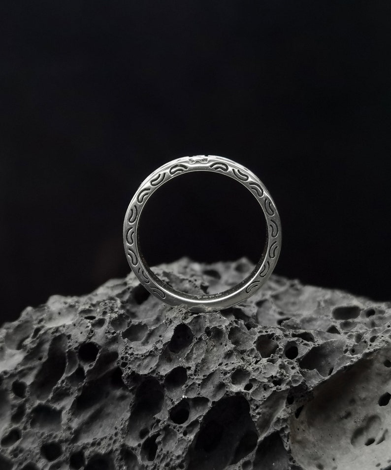 May include: A silver ring with a detailed, intricate design. The ring is sitting on a dark, textured surface.