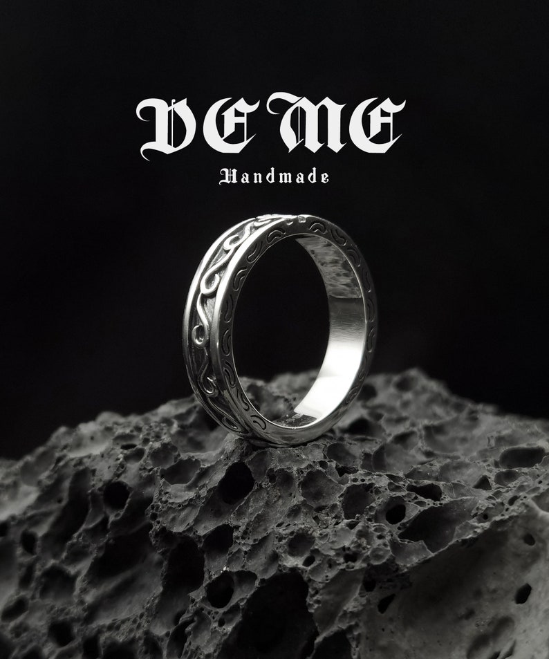 May include: A silver ring with a detailed, intricate design. The ring is handmade and features a decorative band with a wave-like pattern. The ring is sitting on a dark, textured surface.