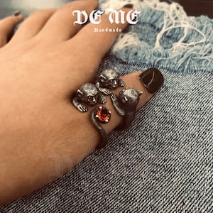May include: Silver ring featuring three monkey figures surrounding a red gemstone. The ring is handmade and the brand name "De Me Handmade" is visible in the background.