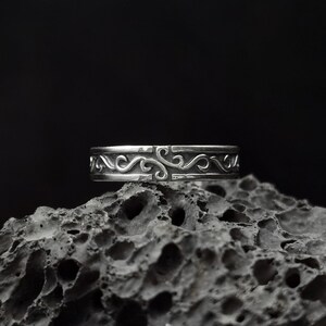 May include: A silver ring with a decorative, swirling design. The ring is sitting on a dark, textured surface.