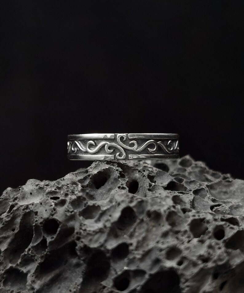 May include: A silver ring with a decorative, swirling design. The ring is sitting on a dark, textured surface.