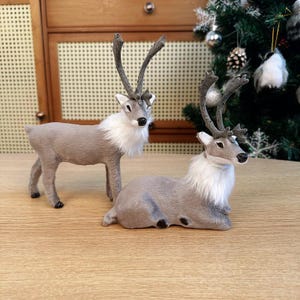 Peut inclure: Deux figurines décoratives de rennes. L'un est debout, l'autre est couché. Les deux ont des corps gris, de la fourrure blanche autour du cou et des bois détaillés. Ils sont posés sur une surface en bois, avec un sapin de Noël et des décorations en arrière-plan.