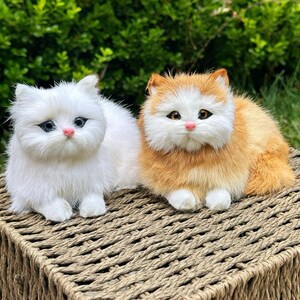 Peut inclure: Deux figurines de chat en peluche, une blanche unie et une orange et blanche, toutes deux dotées de longs poils synthétiques, d'oreilles pointues, d'yeux sombres avec des marques noires et de petits nez roses, côte à côte sur une surface tressée.