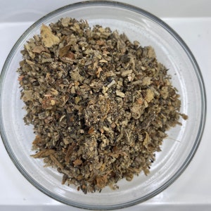 May include: A close-up of a small glass bowl filled with dried herbs. The herbs are a mix of brown, green, and gray.