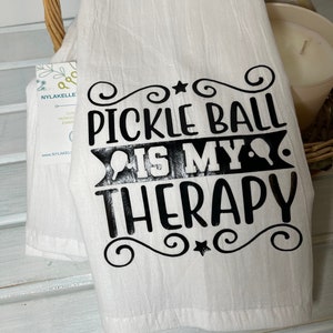 Op de afbeelding: Een witte keukendoek met zwarte tekst die "Pickleball is my therapy" luidt, met een decoratief ontwerp rond de tekst.