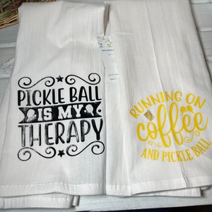 Op de afbeelding: Twee witte keukendoeken met zwarte en gouden tekst. De eerste handdoek zegt "Pickleball is my therapy" en de tweede handdoek zegt "Running on coffee and pickleball".