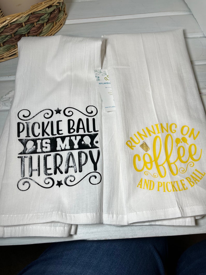 Op de afbeelding: Twee witte keukendoeken met zwarte en gouden tekst. De eerste handdoek zegt "Pickleball is my therapy" en de tweede handdoek zegt "Running on coffee and pickleball".