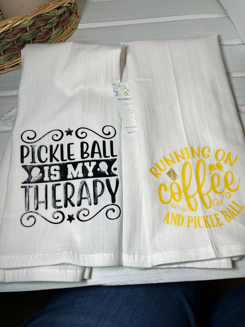 Op de afbeelding: Twee witte keukendoeken met zwarte en gouden tekst. De eerste handdoek zegt "Pickleball is my therapy" en de tweede handdoek zegt "Running on coffee and pickleball".