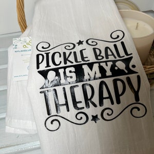 Op de afbeelding: Een witte keukendoek met zwarte tekst die "Pickleball is my therapy" luidt, met een ster en krulletjes rond de tekst.