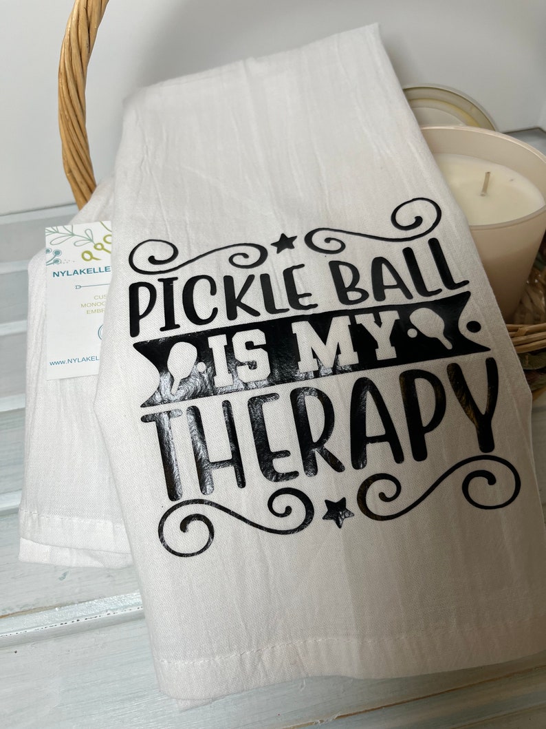 Op de afbeelding: Een witte keukendoek met zwarte tekst die "Pickleball is my therapy" luidt, met een ster en krulletjes rond de tekst.