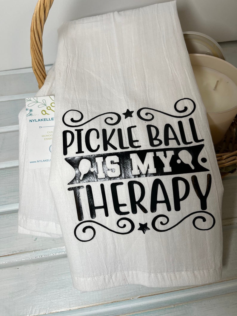 Op de afbeelding: Een witte keukendoek met zwarte tekst die "Pickleball is my therapy" luidt, met een decoratief ontwerp rond de tekst.