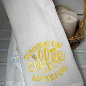 Op de afbeelding: Witte keukendoek met de tekst "Running on coffee and pickle ball" in gouden letters. Een klein glas koffie is afgebeeld in de tekst.