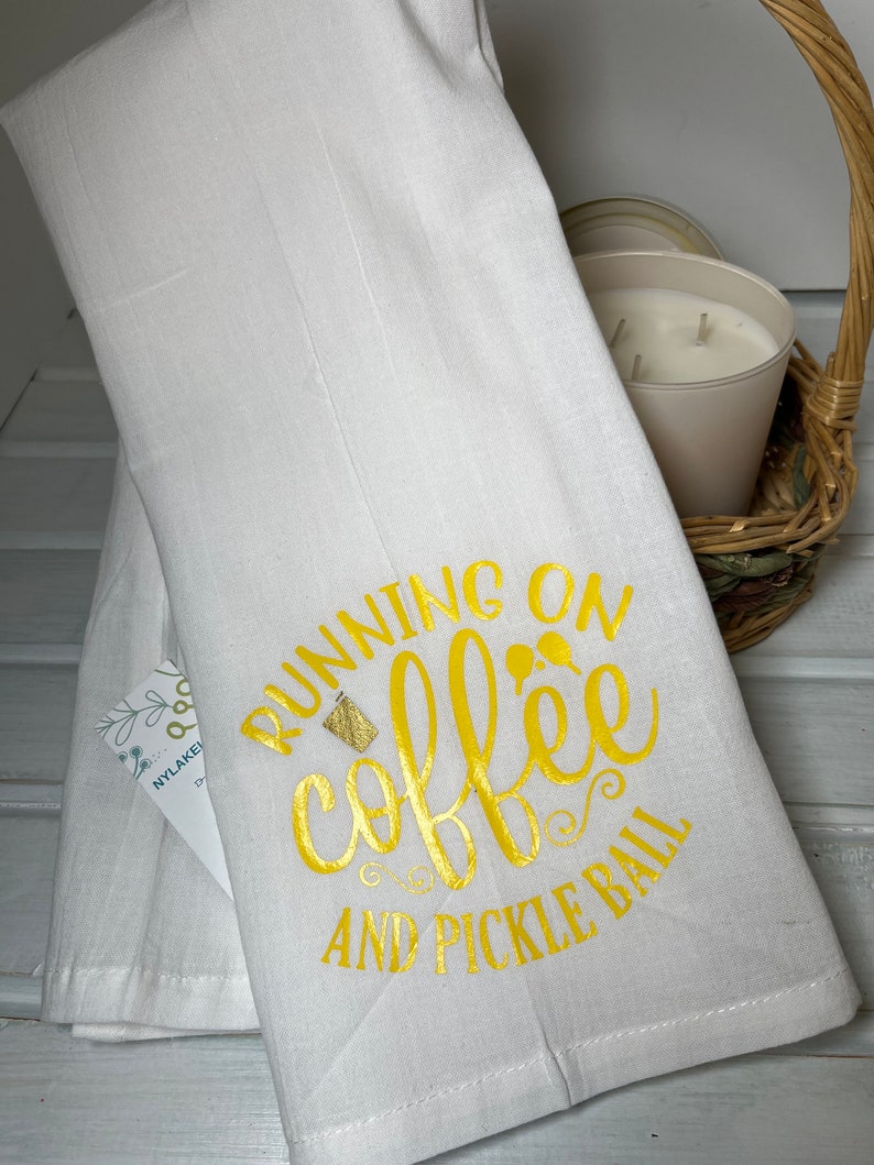 Op de afbeelding: Witte keukendoek met de tekst "Running on coffee and pickle ball" in gouden letters. Een klein glas koffie is afgebeeld in de tekst.