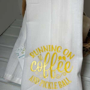 Op de afbeelding: Witte keukendoek met de tekst "Running on coffee and pickle ball" in gouden letters.